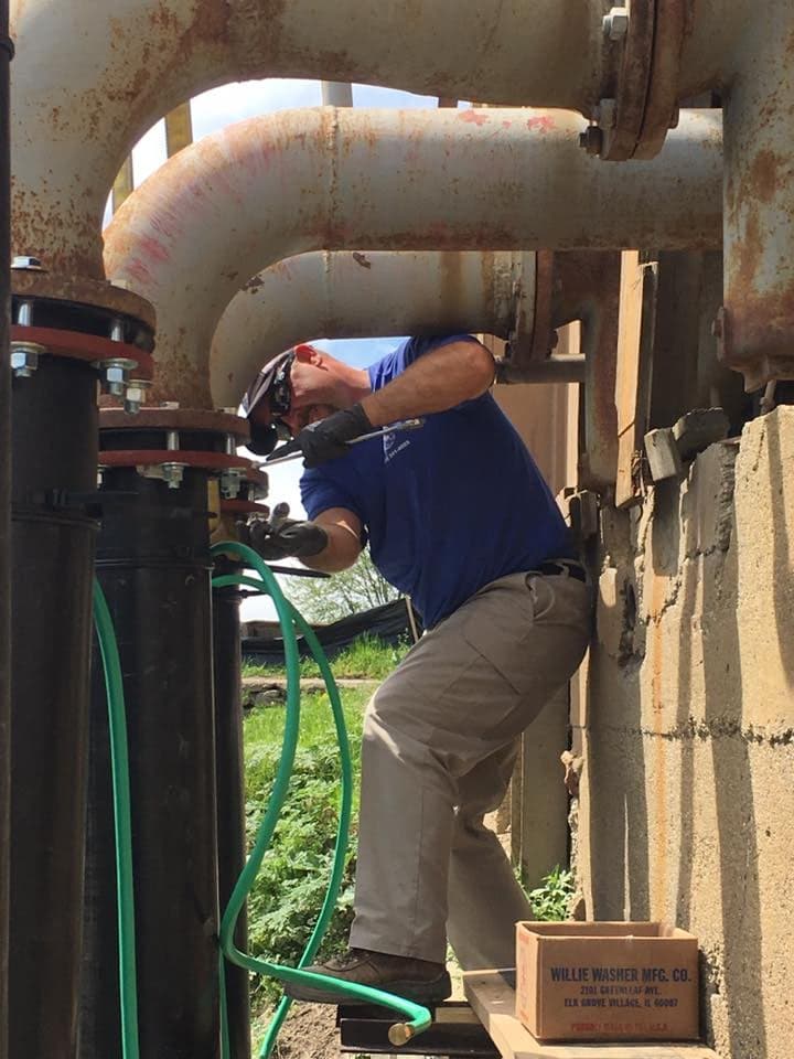 Worker repairing industrial pipes with tools, wearing safety gear on a construction site.