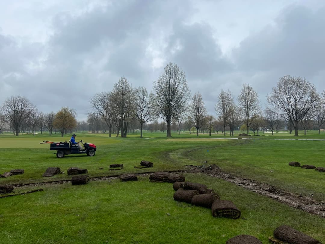 Landscape with a worker on a utility vehicle laying sod on a cloudy day at a golf course.